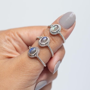 May include: Three silver rings featuring oval-shaped labradorite gemstones with blue and grey iridescence. Each ring has a twisted metal band and a decorative beaded setting surrounding the central stone, showcasing a polished finish.