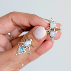 May include: Two silver rings with pear-shaped opal stones. One ring features two opals, while the other has a single pair of opals. The rings are held against a white background, showcasing their intricate designs and iridescent gemstones.