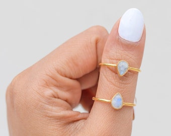 May include: Two gold-colored rings with teardrop-shaped, iridescent gemstones. The rings are displayed on a person's finger against a white background. The gemstones have a subtle blue and white color.