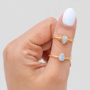 May include: Two gold-colored rings with teardrop-shaped, iridescent gemstones. The rings are displayed on a person's finger against a white background. The gemstones have a subtle blue and white color.