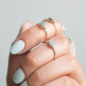 May include: A hand displays several silver rings, each featuring a small, round, opalescent gemstone. The rings have a delicate, minimalist design, with a thin band and subtle decorative elements near the gemstone. The nails are painted a light blue.