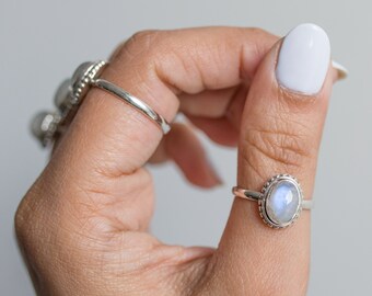 May include: A hand displays several silver rings, including a moonstone ring with an oval-shaped stone. The moonstone has a blue and white iridescent glow. The ring is set in a silver band with a beaded border.