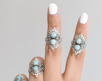 May include: Four silver rings with ornate filigree designs, each featuring two light blue, oval-shaped gemstones. The rings are displayed on fingers against a neutral background, showcasing the jewelry's intricate details and color.
