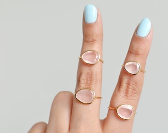May include: Four gold rings with irregular, light pink gemstones. The rings are displayed on fingers with light blue nail polish. The gemstones have a soft, translucent appearance, set in delicate gold bands.