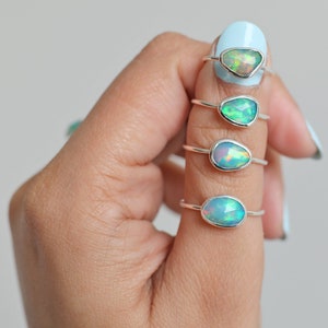 May include: Four silver rings with iridescent opal stones are displayed on a finger. The opals have a range of shapes, including oval and teardrop, and display flashes of green, blue, and orange. The rings are set against a light blue background.