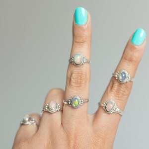 May include: A collection of silver rings, each featuring an oval opal gemstone. The rings are displayed on fingers with turquoise nail polish. The opals display a range of colors, including white, pink, and green.