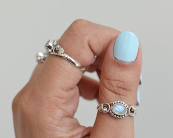 May include: A hand wearing three silver rings. The main ring features an oval-shaped, iridescent gemstone set within a decorative silver band. The other rings have heart-shaped designs and small gemstones.
