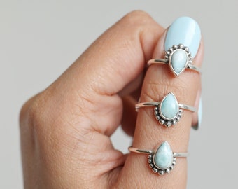 May include: Three silver rings with teardrop-shaped, light blue gemstones. Each ring features a beaded design around the gemstone. The rings are displayed on a light background.
