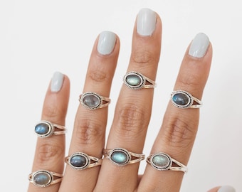 May include: A hand displays several silver rings, each featuring an oval, iridescent gemstone. The rings have a split-shank design and a rope-like border around the stone. The gemstones display shades of blue, gray, and green.