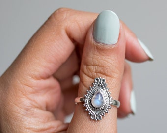 May include: A silver ring with a teardrop-shaped moonstone. The ring features an ornate, filigree design around the stone, with a detailed border. The moonstone has a pearly, iridescent appearance. The ring is worn on a finger.