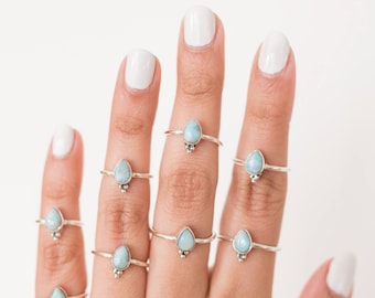 May include: A hand displays several silver rings, each featuring a light blue, teardrop-shaped gemstone. The rings are set on a thin band with a small decorative element at the base of the gemstone. The background is plain white.