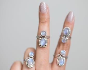 May include: Several sterling silver rings with oval and round moonstone cabochons are displayed. Each ring features a larger oval stone and a smaller round stone, set on a delicate band. The moonstones have a soft, milky blue color.