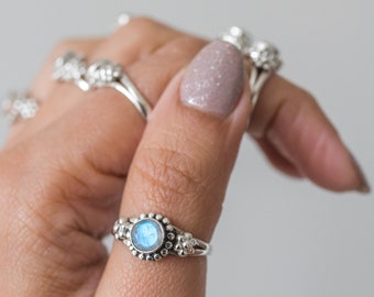 May include: A silver ring with a round, light blue gemstone center, surrounded by small silver beads and floral accents. The ring is on a finger, with other rings visible in the background. The ring's design is intricate and delicate.