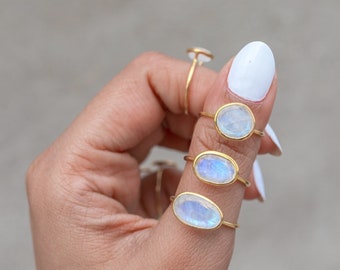 May include: Several gold rings with oval, iridescent moonstone settings are displayed on a hand. The rings have a delicate, thin band. The moonstones have a blue and white color. The background is a neutral gray.