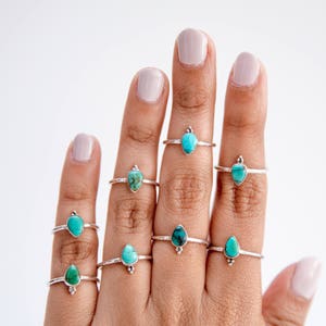 May include: A hand displaying several silver rings with teardrop-shaped turquoise stones. Each ring has a delicate band and small silver accents near the stone. The turquoise stones vary in shades of blue and green.