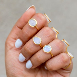K&ouml;nnte beinhalten: Eine Hand zeigt mehrere goldfarbene Ringe, jeder mit einem hellfarbigen, ovalen Stein. Die Ringe haben ein schlichtes, elegantes Design, wobei der Stein in einer d&uuml;nnen Goldfassung gefasst ist. Die N&auml;gel sind wei&szlig; lackiert.