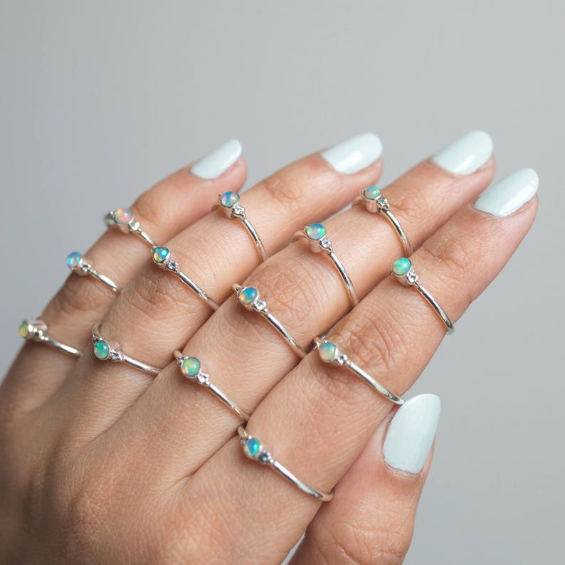 May include: A collection of silver rings, each featuring a small, round opal gemstone. The rings are displayed on a hand with light blue nail polish. The opals have a mix of colors, including blue, green, and white.