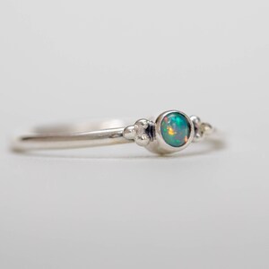 May include: A delicate silver ring featuring a round, iridescent opal gemstone. The ring has a thin band with small, decorative bead accents on either side of the opal. The opal displays a mix of green, blue, and orange hues.