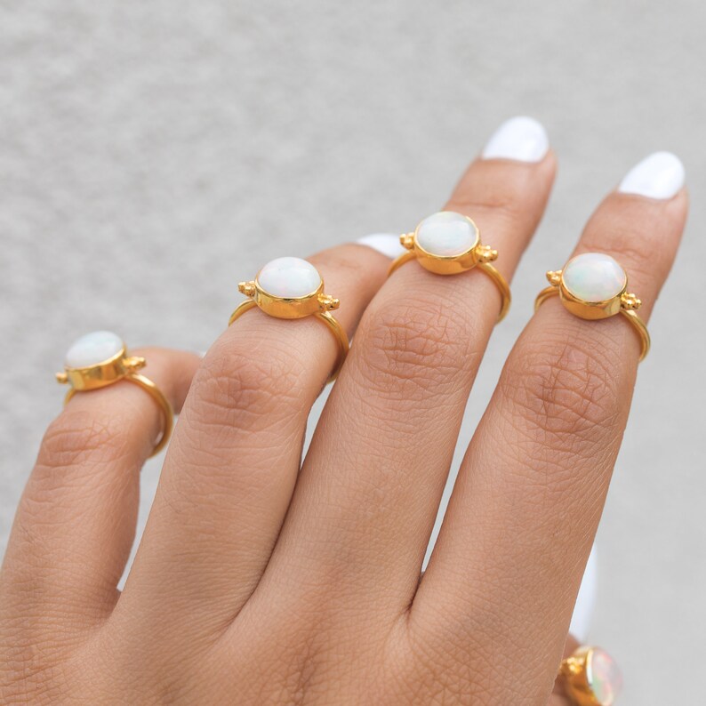May include: A hand displays several gold rings, each featuring a round, white gemstone. The rings have a simple band and decorative accents on either side of the gemstone. The rings are displayed against a neutral background.