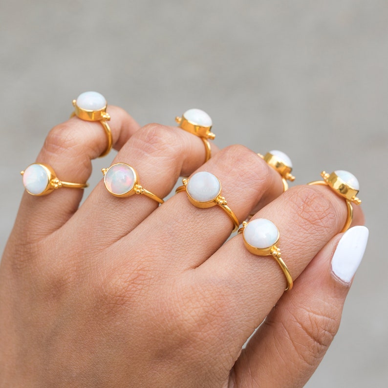 May include: A hand displays several gold rings, each featuring a round, white opal stone. The rings have a delicate, thin band and are set in a gold bezel. The opal stones have a pearly, iridescent sheen.