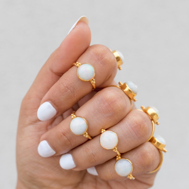 May include: A collection of gold rings, each featuring a large, round, white opal stone. The rings have a delicate, thin band and small decorative accents. The rings are displayed on a hand with white nail polish.