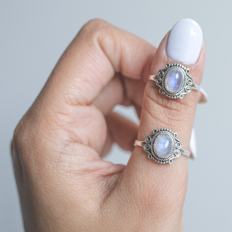 May include: Two silver rings with oval, iridescent moonstone cabochons. The rings have ornate, filigree-style settings. The rings are displayed on a person's finger against a white background. The rings are a type of jewelry.