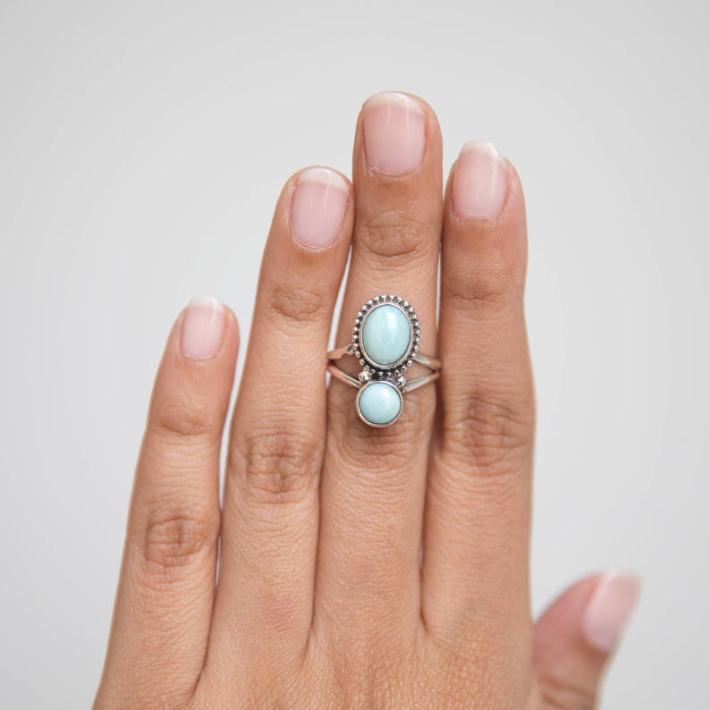 May include: A sterling silver ring featuring two oval-shaped, light blue gemstones. The top gemstone is larger and surrounded by a beaded border. The ring is displayed on a hand against a white background, showcasing the ring's design and color.