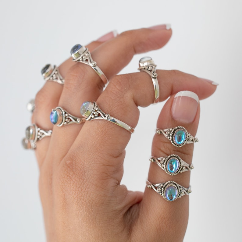Pu&ograve; includere: Una collezione di anelli d'argento con pietre preziose ovali e iridescenti. Gli anelli hanno fasce decorative e dettagliate. Le pietre preziose riflettono diversi colori. Mostrati su una mano.