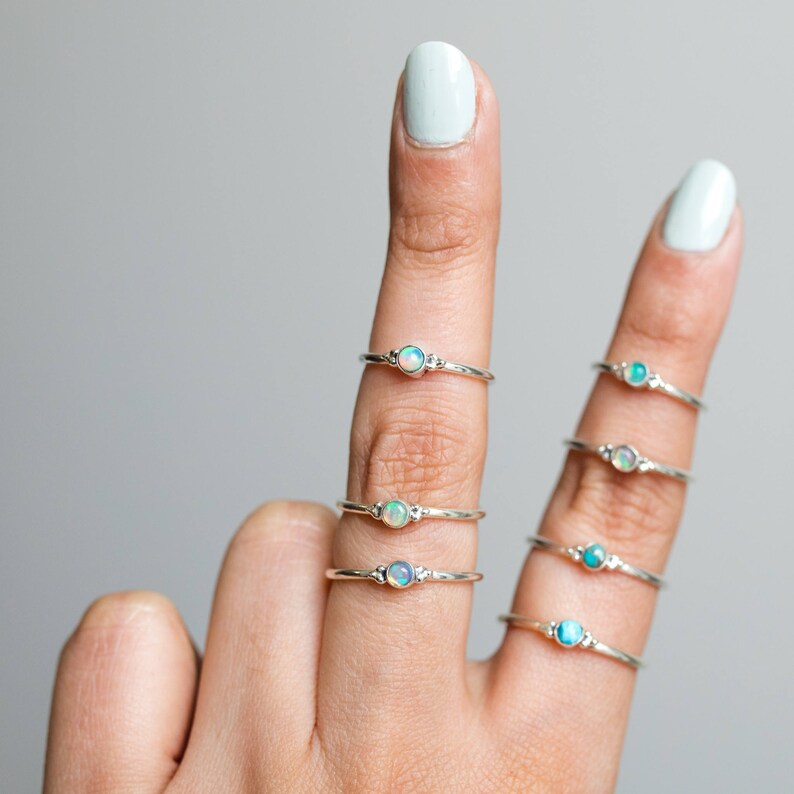 May include: Several silver rings with small, round opal stones are displayed on fingers. The rings have a delicate band and a simple design, with the opal as the focal point. The opals have a mix of white, blue, and green hues.
