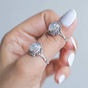 May include: Two silver rings with oval, iridescent moonstone cabochons. The rings have ornate, filigree-style settings. One ring is on a finger, and the other is nearby. The background is a soft, neutral color.