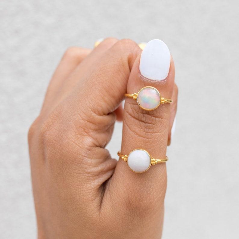 May include: Two gold rings with round, white gemstones. One ring features an iridescent opal, the other a solid white stone. The rings are set on delicate gold bands, showcasing a minimalist design. The rings are displayed on a hand with white nail polish.
