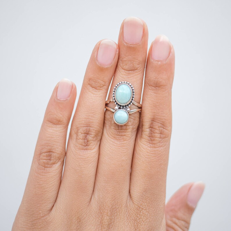 May include: A sterling silver ring with two oval, light blue gemstones. The top gemstone is larger and surrounded by a beaded border. The ring is on a hand against a white background.