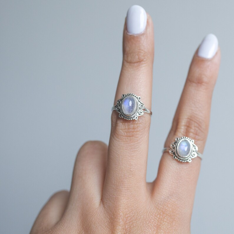 May include: Two silver rings with oval, iridescent moonstone cabochons. The rings have ornate, filigree-style settings. One ring is on a finger, and the other is in the background. The background is a solid, light gray.