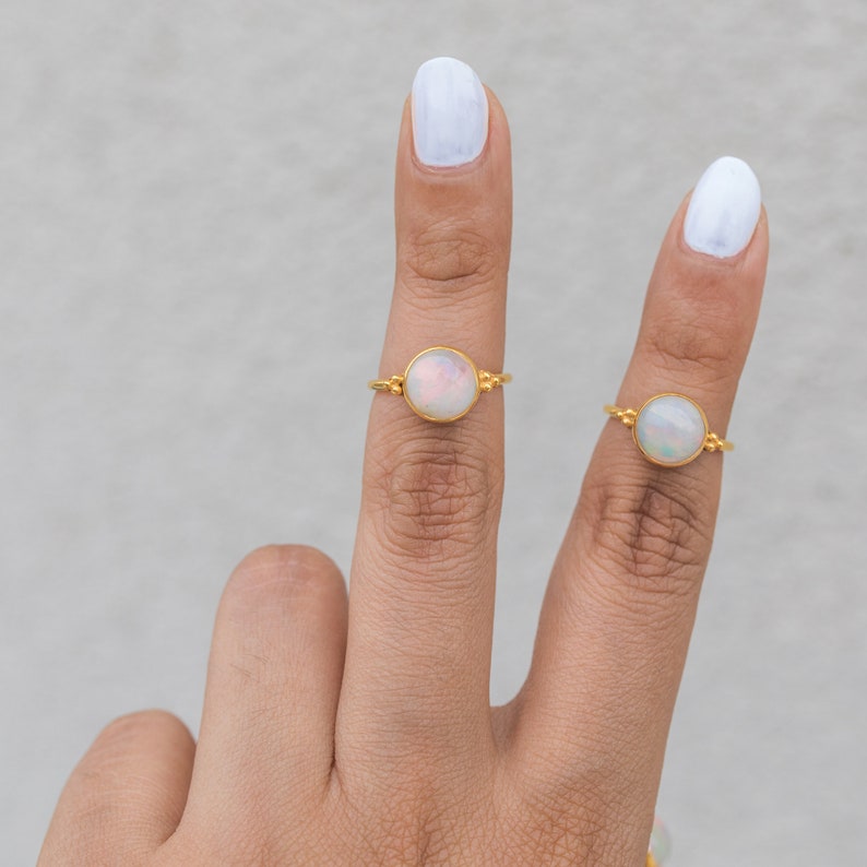 May include: Two gold rings, each featuring a round, iridescent opal stone. The rings are displayed on fingers with white nail polish against a neutral background. The gold bands have small decorative details on either side of the opal.