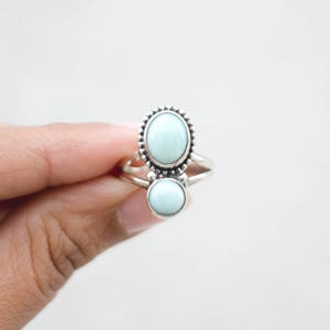 May include: A silver ring featuring two light blue gemstones. The top stone is oval-shaped and surrounded by a beaded border. The bottom stone is round. The ring is held by a hand against a white background.