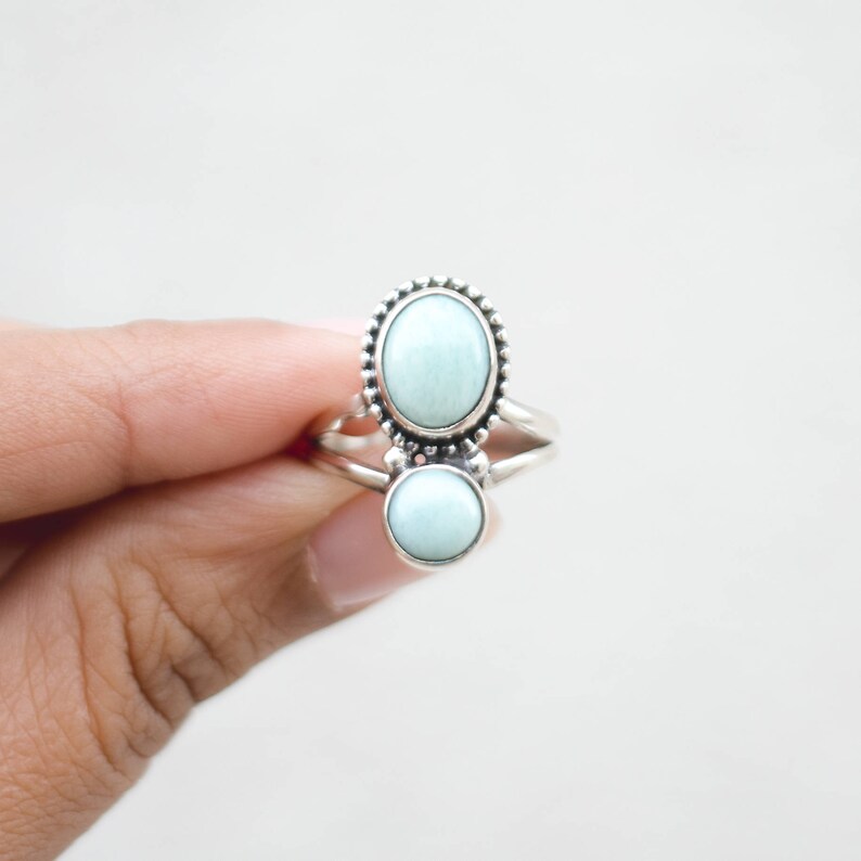 May include: A silver ring featuring two light blue gemstones. The top stone is oval-shaped and surrounded by a beaded border. The bottom stone is round. The ring is held by a hand against a white background.