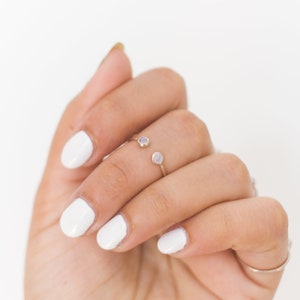 May include: A gold-colored ring with two small, round, light-colored gemstones. The ring is worn on a hand with white nail polish. The ring's design features an open band with a gemstone on each end.