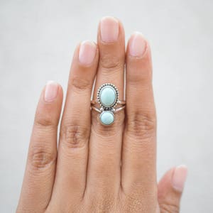 May include: A silver ring set with two oval and round, light blue gemstones. The larger stone is surrounded by a beaded border. The ring is displayed on a hand against a white background.