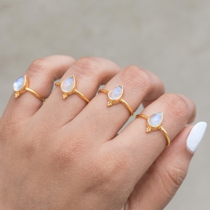 May include: Four gold rings, each featuring a pear-shaped, iridescent gemstone. The rings are displayed on a hand, showcasing the delicate design and the play of light on the stones. The rings have a small decorative element at the base of the gemstone.