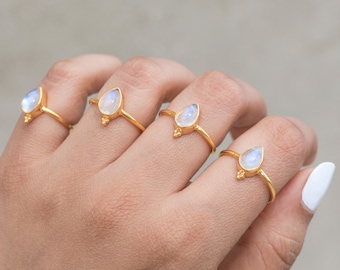 May include: Four gold rings, each featuring a pear-shaped, iridescent gemstone. The rings are displayed on a hand, showcasing the delicate design and the play of light on the stones. The rings have a small decorative element at the base of the gemstone.