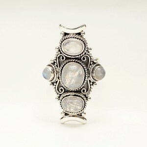 Peut inclure: Une bague argent&eacute;e orn&eacute;e de cinq cabochons en pierre de lune ovales et ronds. La bague pr&eacute;sente un design orn&eacute; avec des d&eacute;tails en forme de corde autour des pierres et des ornements d&eacute;coratifs. Les pierres de lune ont un aspect laiteux et iris&eacute;.