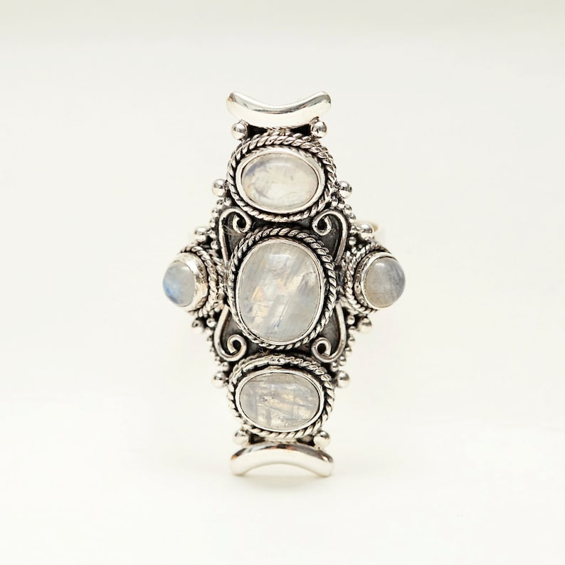 Peut inclure: Une bague argent&eacute;e orn&eacute;e de cinq cabochons en pierre de lune ovales et ronds. La bague pr&eacute;sente un design orn&eacute; avec des d&eacute;tails en forme de corde autour des pierres et des ornements d&eacute;coratifs. Les pierres de lune ont un aspect laiteux et iris&eacute;.