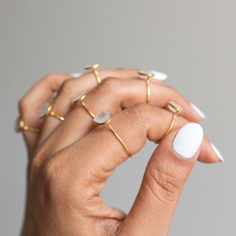 K&ouml;nnte beinhalten: Eine Sammlung goldfarbener Ringe, jeder mit einem kleinen, runden, irisierenden Stein. Die Ringe werden an einer Hand mit wei&szlig;em Nagellack gezeigt. Das minimalistische Design der Ringe wird vor einem neutralen Hintergrund hervorgehoben.