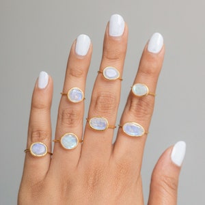 K&ouml;nnte beinhalten: Eine Auswahl goldfarbener Ringe, jeder mit einem einzigartigen, irisierenden, ovalen oder runden Edelstein. Die Ringe werden auf einer Hand mit wei&szlig;em Nagellack vor einem neutralgrauen Hintergrund pr&auml;sentiert. Die Edelsteine haben ein milchiges, opalisierendes Aussehen.
