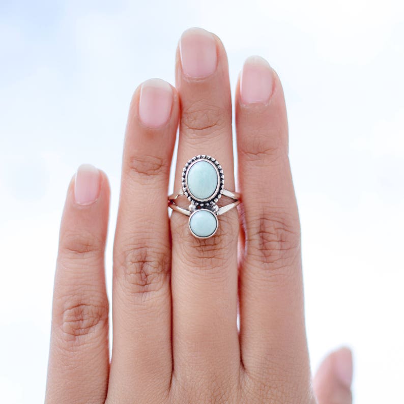 May include: A silver ring featuring two oval-shaped, light blue gemstones. The top stone is larger and surrounded by a beaded border. The ring is on a finger against a light background.