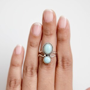 May include: Two silver rings with oval and round, light blue gemstones. The top ring has a beaded border around the oval stone. The rings are stacked on a finger against a white background.