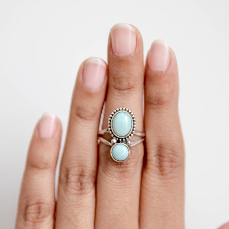 May include: Two silver rings with oval and round, light blue gemstones. The top ring has a beaded border around the oval stone. The rings are stacked on a finger against a white background.