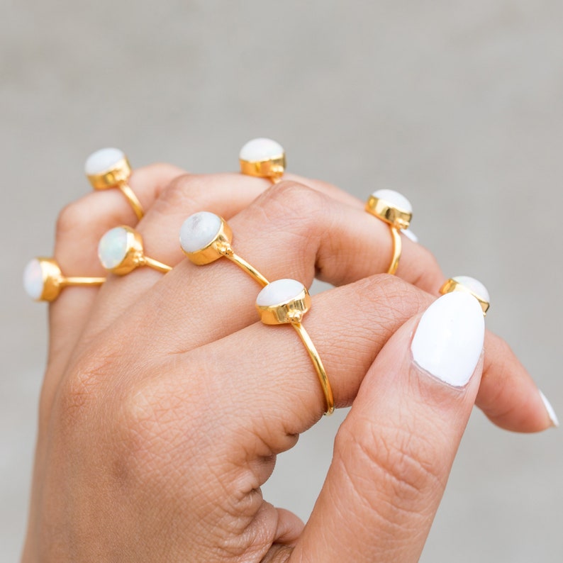 May include: A collection of gold-colored rings, each featuring a round, white gemstone. The rings are displayed on a hand with white nail polish. The rings have a minimalist design, with a thin band and a prominent gemstone.