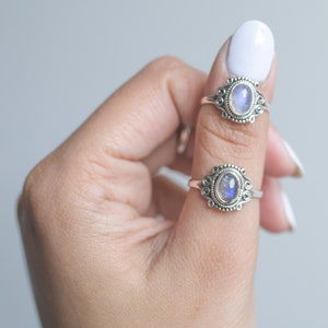 May include: Two silver rings with oval, iridescent gemstones are displayed on a finger. The rings have ornate, filigree-style settings. The gemstones have a blue and white color. The background is a soft, neutral gray.