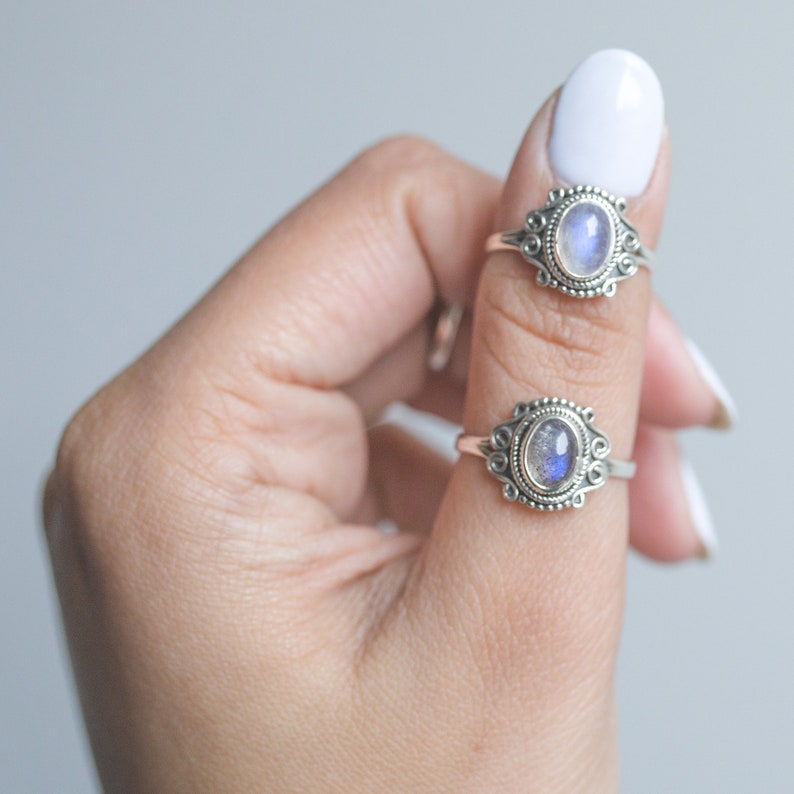 May include: Two silver rings with oval, iridescent gemstones are displayed on a finger. The rings have ornate, filigree-style settings. The gemstones have a blue and white color. The background is a soft, neutral gray.